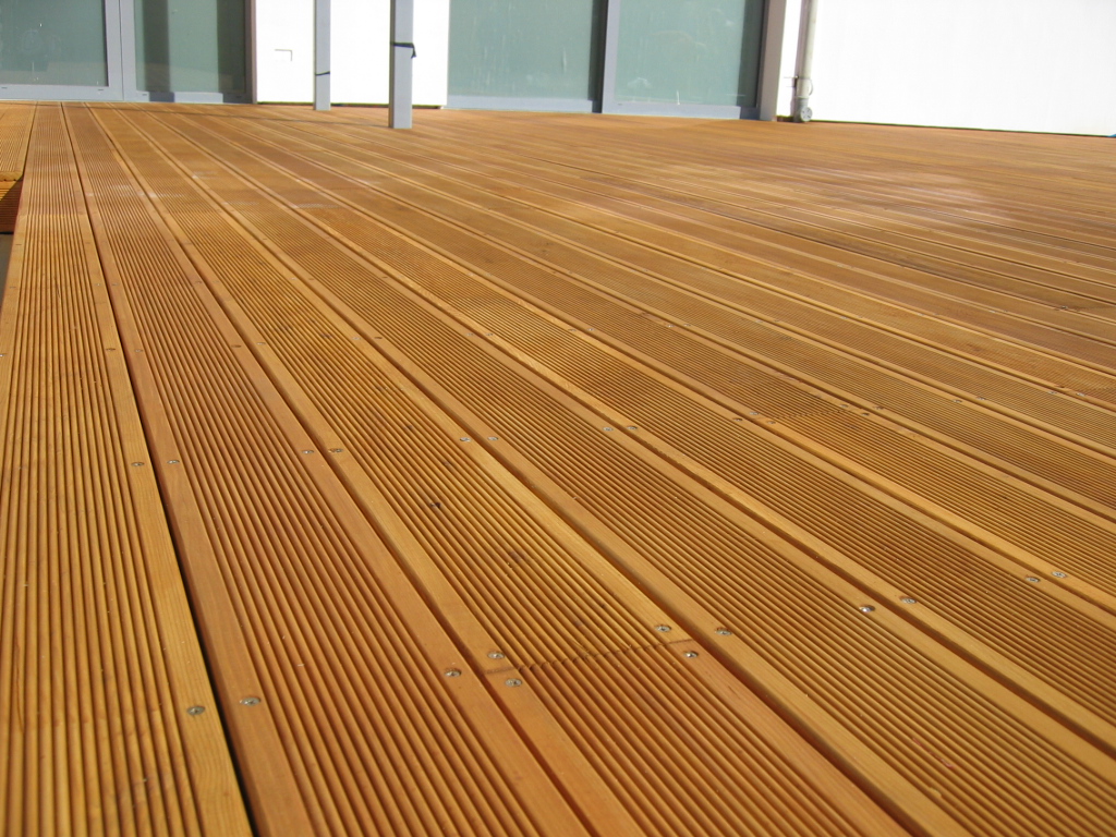 Nowy taras z deski kompozytowej z modrzewia syberyjskiego, widoczne ryflowanie i mocowania, z białymi ścianami i przeszklonymi drzwiami w tle.